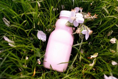 Damlacıklı pembe probiyotik içecek şişesi, çiçek yapraklarıyla taze çimlerin üzerinde yatıyor. Paket tasarımı ve ürün sunumu için ideal model.