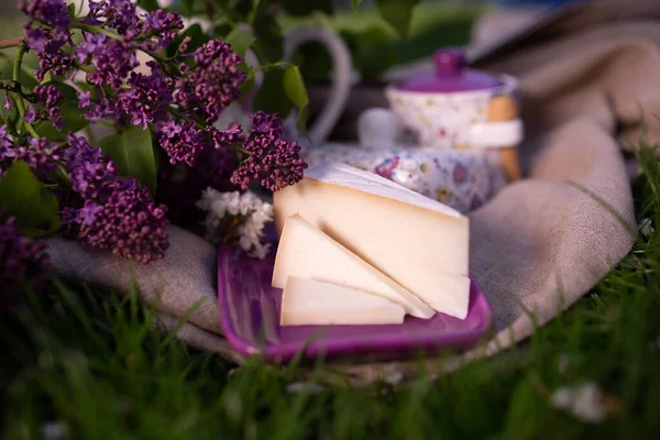 Mor bir tabakta servis edilen sert beyaz peynir dilimleri leylak çiçekleri ve kırsal kumaş, açık hava bahar piknik seti..