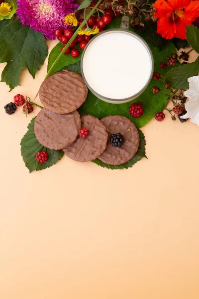 Yeşil yapraklar ve renkli çiçeklerle süslenmiş bir fincan sütün yanında böğürtlenli kurabiye manzarası..