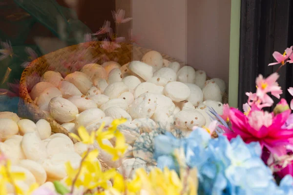 28 Ağustos 2025, Bruges, Belçika. Bir dükkan vitrininde renkli çiçeklerle çevrili susam tohumları serpiştirilmiş küçük, yuvarlak, beyaz bir sepet..