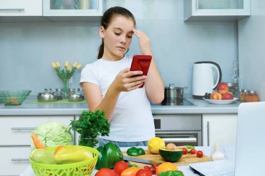 Mutfakta yemek yapan ve telefonda yemek tarifi arayan güzel bir genç kız. Dizüstü bilgisayar mutfak masasında. Gıda blogcusu kavramı
