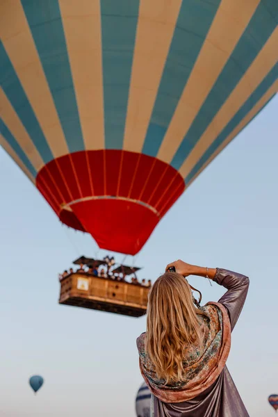 Beautiful blonde woman photographer takes pictures of flying hot air balloon at dawn in Cappadocia, Turkey. Travel concept.  Dressed in a scarf with a Turkish national pattern Ebru