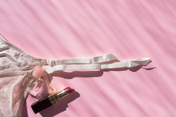 Lingerie garter for stockings, perfume bottle, lipstick and pearl beads. Pink flatlay top view with place for text. Picture with sunbeams. Taking care of yourself. Self-love. Fashionable modern girl stuff