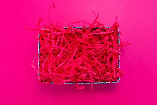 Pink shredded paper packaging material in box flat lay on pink background. Template for placing goods or text