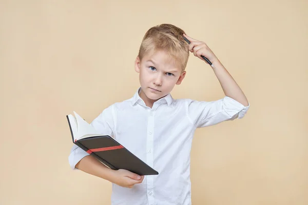 Sevimli akıllı çocuk, düşünceli ve elinde bir defter tutarken kalemle başını kaşıyor..