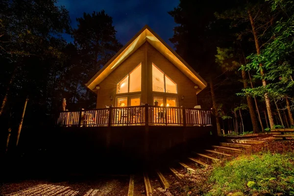 森の中の照明付きの窓で休暇の家 夜に撮影された牧歌的なキャビンやコテージ 概念には デザイン 屋外生活などがあります ロイヤリティフリーのストック画像