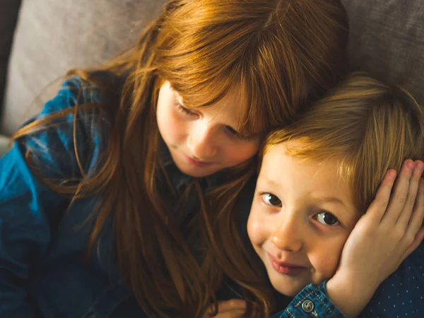 İki sarışın kardeş çocuklarda bir kanepe sarılma üzerinde mavi gömlek. Mutlu sevimli çocuklar kapalı portresi. Portre