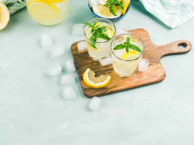 Taze limonata, buzlu bardaklar, nane yaprakları ve ahşap tahtada parlak gri ve mavi arka planda limon dilimleri.