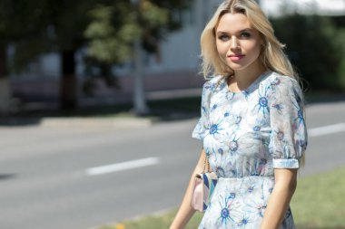 Açık mavi elbiseli genç bir kız, güneşli bir yaz gününde bulanık bir arkaplanda utançla sokakta yürür.