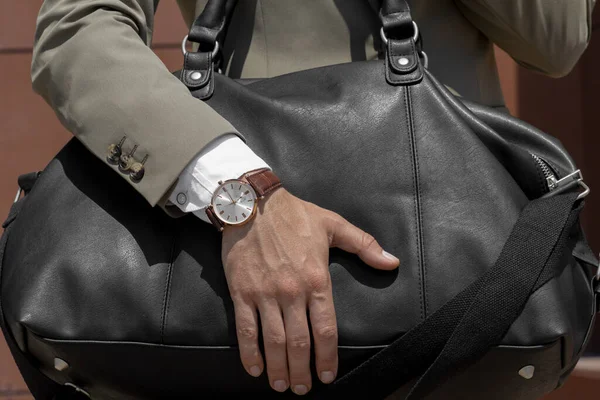 A man in a suit holds with his hand on which a quartz watch is dressed, a travel bag with a valuable load