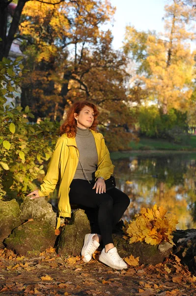 young woman in a yellow jacket on a yellow autumn background sitting in the Park on the stones