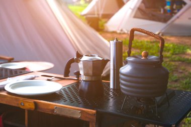 Klasik açık hava çaydanlığı, kahve öğütücü ve açık mutfak ekipmanları kamp masasında güneş ışığı ile birlikte doğal park alanındaki çadırların yüzeyinde.