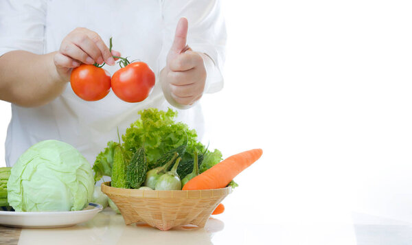 Cropped image of fat woman holding fresh tomatoes and showing thumps up with non-toxic vegetables in a bamboo basket on marble table in white kitchen, healthy food for overweight people concept