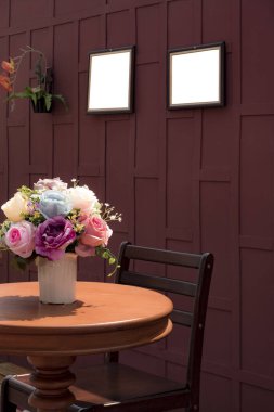 Tahta ahşap koltuklu ahşap masa üzerinde beyaz seramik vazoda suni gül çiçeklerinin yan görüntüsü ve ahşap duvarda eski tarz ve dikey çerçeveli boş resim çerçevesi