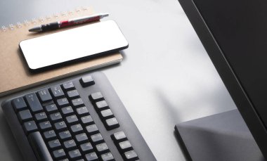 Akıllı telefonun yüzeyindeki sabah ışığı, tükenmez kalem, bilgisayar klavyesinin bir parçasına sahip spiral defter ve gri masa üstünde monitör, yüksek açılı görüş ve seçici odak noktası oluşturur.