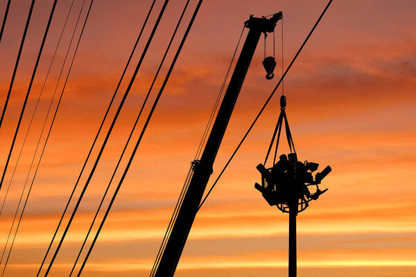 Silhouette low angle view of mobile crane lifting electrician in telescopic boom to install electrical system on spotlights tower against colorful sunset sky background