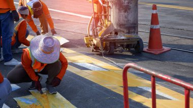 Yansıtıcı yelekli yol işçileri, termoplastik sprey yol işaretleme makinesiyle asfalt yolda trafik sarı çizgilerini boyamak için çalışıyorlar. Demiryolu rayları cadde yüzeyinde kesişiyor, seçici odaklı.