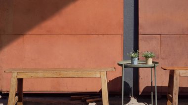 Boş ahşap banklar ve binaların dışındaki dinlenme yerlerinde güneş ışığı ve gölgeli kayan duvarın önündeki yuvarlak metal masadaki saksılarda bulunan iki küçük yeşil bitki.  