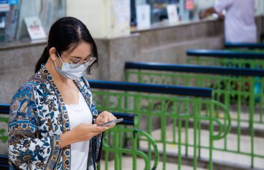 Tren istasyonundaki bilet gişelerinde akıllı telefon kullanan sıradan görünümlü, koruyucu maskeli genç Asyalı kadın turistler.