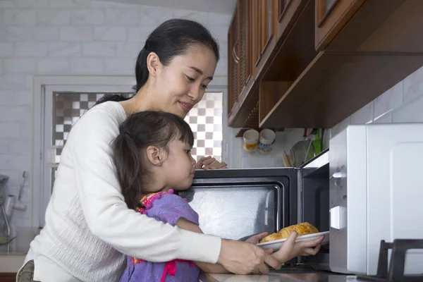 Asian mother is teaching her little daughter to use a microwave oven to warm croissants in the kitchen at home