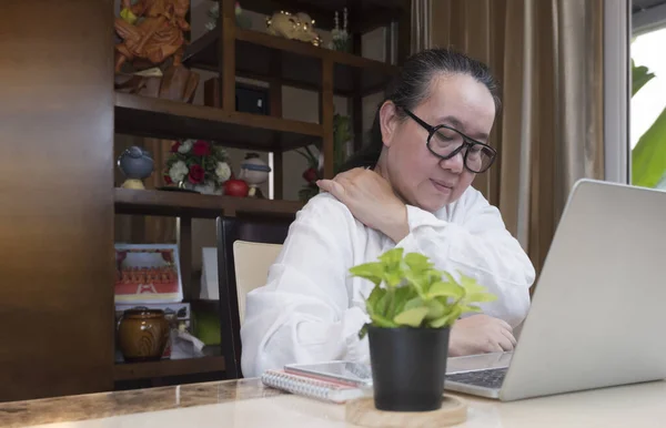 Asyalı olgun bir kadının uzun süre evde oturma odasında dizüstü bilgisayarla çalışırken omzunu sıktığını görüyorum.