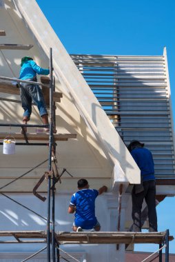 İskele üzerinde çalışma araçları olan Asyalı inşaat işçileri, eski ev binasının dış çatı yapısını dikey çerçevede yenilemek için çalışıyorlar