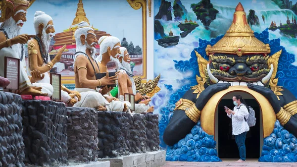 Asian female tourist in protective mask using smartphone to search information while walking to look Buddhist art statues around Thai temple area
