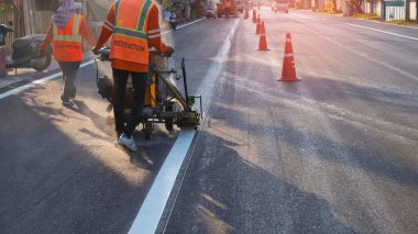 Gece vakti güneş ışığı ve gölgeyle asfalt yol yüzeyindeki trafiği beyaza boyamak için çalışan termoplastik sprey işaretleme makinesiyle yol işçilerinin arka görüntüsü, seçici odaklanma