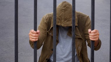 Kapüşonlu ümitsiz adam başını eğip demir çubuklar tutuyor, göçmen ve mülteci krizi umutsuz bir konsept içinde.