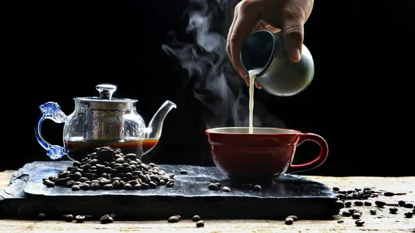 Man\'s hand pouring milk into red  coffee cup with steam, coffee beans and coffee pot on black slate with wooden table in vintage dark tone style