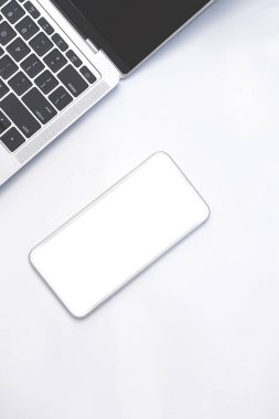 Beyaz akıllı telefon modeli, boş ekran ve siyah dizüstü bilgisayarın bir kısmı beyaz masanın üstünde dikey çerçevede.