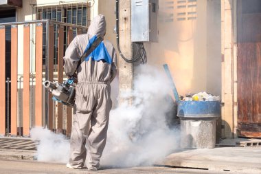 Healthcare worker using fogging machine spraying chemical to eliminate mosquitoes in drainage ditch between two houses.