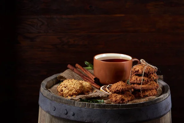 Conceptual festive composition with a cup of hot tea, a large set of cookies, cinnamon sticks bunch and dried oranges on a wooden barrel over rustic background, selective focus, close-up. Delicious