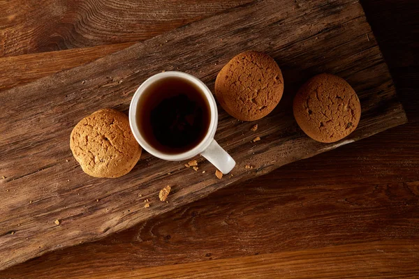 Білий порцеляновий кухоль чаю та солодке печиво на шматочку дерева на дерев'яному фоні, вид зверху, вибірковий фокус — стокове фото
