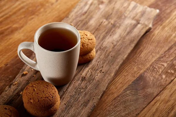 White porcelain mug of tea and sweet cookies on piece of wood over wooden background, top view, selective focus — Stock Photo, Image