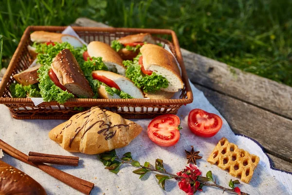 Cesta de picnic llena de sándwiches, baguette y croissant en un mantel hecho en casa, puesta plana, enfoque selectivo — Foto de Stock