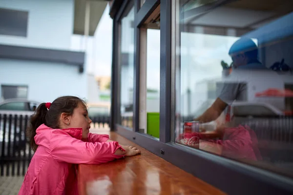 Περίεργος κοριτσάκι περιμένει το burger shop Οδός παράθυρο. — Φωτογραφία Αρχείου