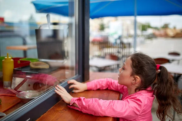 거리 상점 창에서 그녀의 햄버거를 기다리는 호기심 어린 소녀. — 스톡 사진