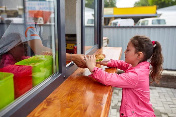 Счастливая маленькая девочка выносит свой бургер через окно уличного магазина . — стоковое фото