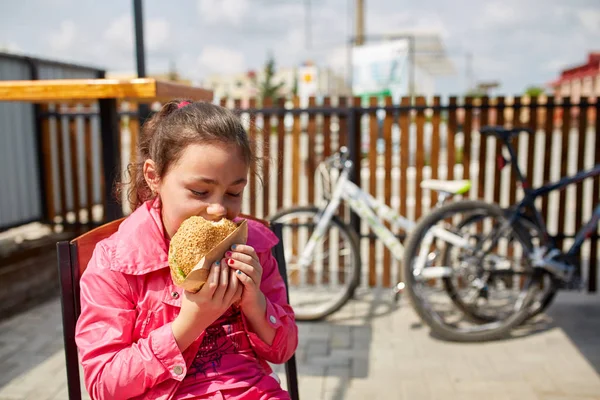 Uma menina está comendo um hambúrguer de queijo no café ao ar livre na frente do estacionamento da bicicleta . — Fotografia de Stock