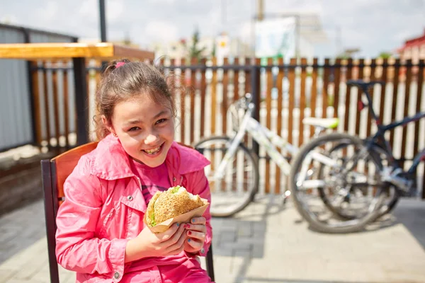 Uma menina está comendo um hambúrguer de queijo no café ao ar livre na frente do estacionamento da bicicleta . — Fotografia de Stock