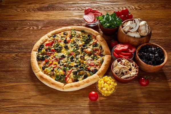 Pizza bodegón. Pizza recién horneada y sus componentes dispuestos sobre fondo de madera . — Foto de Stock