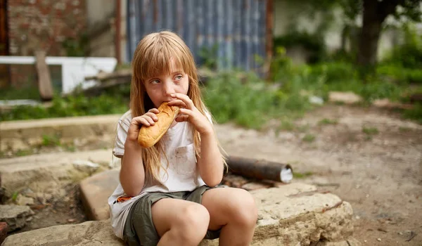 Бедная бездомная девочка ест хлеб в грязном переулке. — стоковое фото