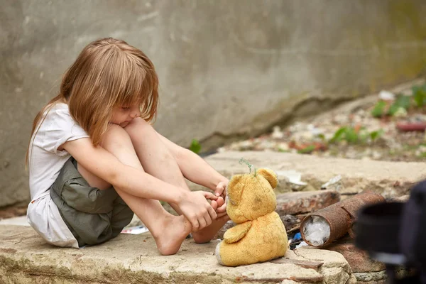 Wees met blote voeten in een armoedige kleren spelen met gebruikt teddybeer op straat. Stockfoto