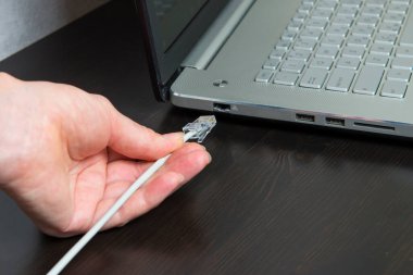 Adamın eli kabloyu siyah masanın üzerinde duran laptopa bağlıyor.