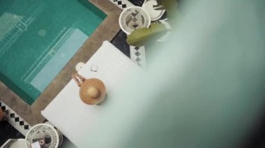 Yukarıdaki görüntülerde geniş kenarlı şapkalı bir kadın havuz kenarında bir yatağın üzerinde oturuyor.