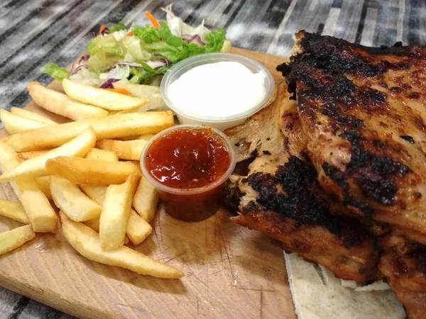Chicken grill, french fries, chili sauce, mayonnaise, and salad served on a wooden plate.