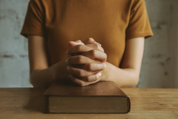 Woman with Bible praying, hands clasped together on her Bible on wooden table.