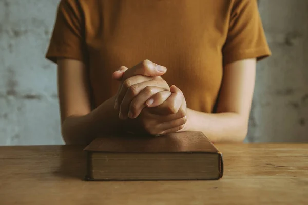 Woman with Bible praying, hands clasped together on her Bible on wooden table.