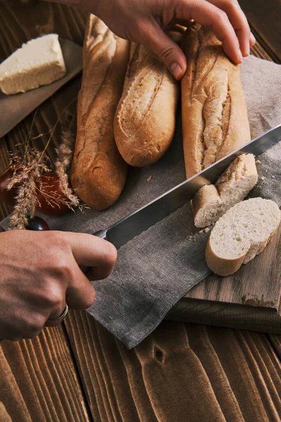 Feche as mãos cortando a baguete francesa fresca em um pano de linho em uma mesa de madeira — Fotografia de Stock